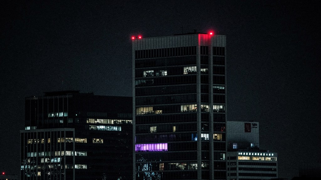 Night view of a financial district skyline through rain-streaked glass, a red warning light blinking atop the tallest tower