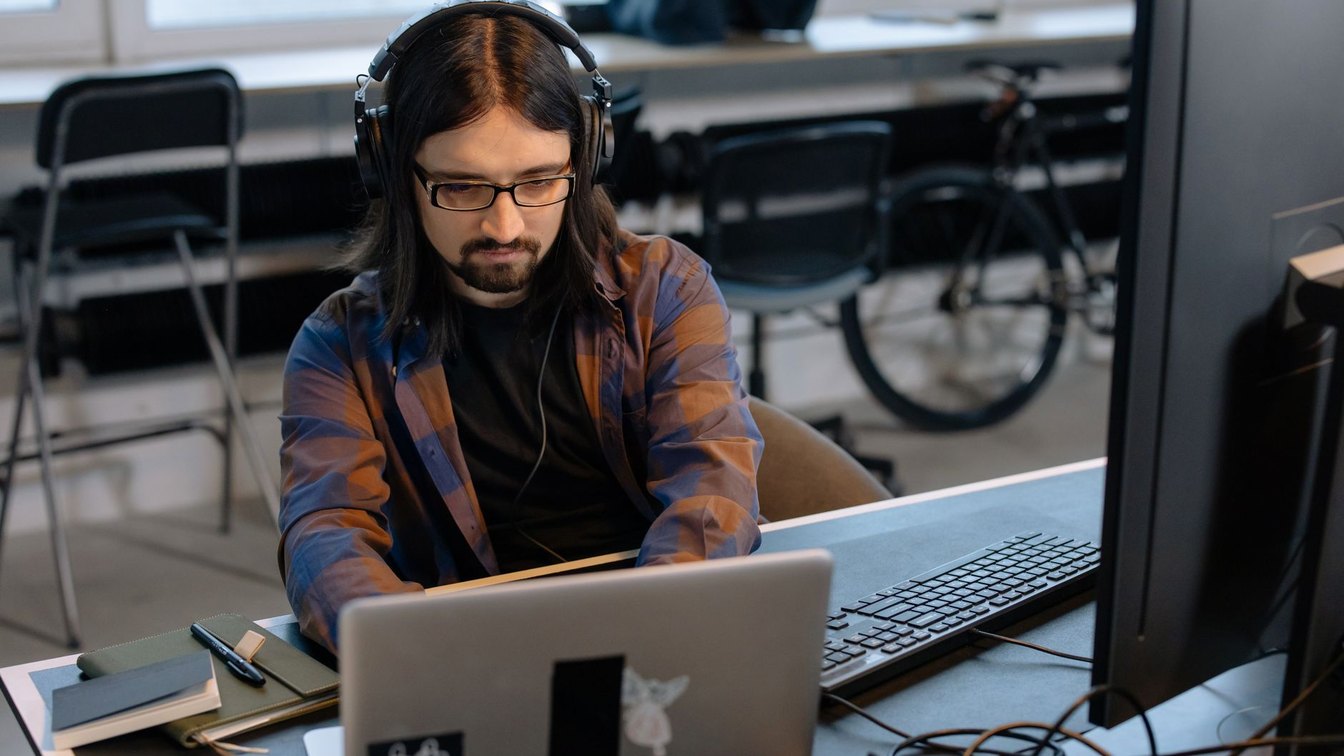 Bearded male developer wearing headphones, coding on a laptop in a modern office setting